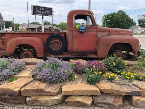 A rusty red vintage pickup truck is parked as a display behind a stone garden wall with purple and yellow flowers, possibly at a Flea market near Coffeyville KS.